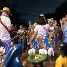 Integrantes de Folia de Reis do Sertão Carioca se reúnem ao ar livre à noite, vestidos com roupas coloridas, chapéus e instrumentos musicais como violas e pandeiros. Em primeiro plano, um presépio artesanal com figuras de barro representa o nascimento de Jesus. Ao fundo, o grupo canta e toca em celebração tradicional, marcada pela devoção, alegria e forte expressão da cultura popular.