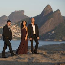 O trio musical T’RIO posa sobre uma formação rochosa à beira-mar, com o Morro Dois Irmãos e o Vidigal ao fundo, banhados pela luz suave do entardecer. Os três integrantes estão elegantes: a mulher veste um longo vestido vinho, enquanto os dois homens usam ternos escuros. A imagem transmite sofisticação e harmonia, refletindo o estilo refinado do grupo.