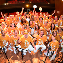 A Companhia Musical Rio Pandeiro reúne-se no palco para uma foto vibrante e cheia de energia. O grande grupo, vestido com camisetas laranja comemorativas, sorri, ergue instrumentos e faz poses animadas. Pandeiros, tambores e outros instrumentos de percussão destacam-se na cena. Ao fundo, veem-se as poltronas vermelhas do teatro, reforçando o clima de celebração e espetáculo coletivo.