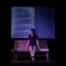 A foto mostra uma mulher sentada em um banco de madeira, em um ambiente de iluminação cênica em tons de roxo e azul. Ela usa um vestido escuro e olha para o lado, com expressão pensativa. Ao fundo, vê-se um painel desfocado que reforça o clima teatral da cena.