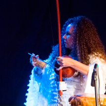 Mulher adulta, identificada como a cantora, compositora, berimbalista e ativista cultural Zilá Lima, aparece em apresentação sobre um fundo escuro. Ela tem cabelos cacheados volumosos e veste roupa clara com textura leve. Zilá toca um berimbau com concentração, iluminada por luz azulada que destaca seus movimentos e a atmosfera cênica da performance.