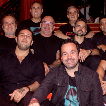 Grupo de nove homens, integrantes da banda de Rock ’n’ Roll Cadillac, está reunido em uma escada de ambiente interno, posando para a foto. Todos vestem roupas escuras e sorriem para a câmera. A iluminação quente destaca os rostos e o corrimão ornamentado ao fundo, criando uma atmosfera descontraída e vibrante, típica do universo do rock.