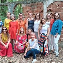 Integrantes do Acorde Vocal, grupo vocal feminino, posam juntas ao ar livre diante de uma antiga parede de tijolos aparentes. As mulheres vestem roupas coloridas e variadas, algumas em pé e outras agachadas, sorrindo para a câmera. O cenário natural, com folhas e vegetação ao redor, transmite acolhimento, união e a força coletiva do grupo.