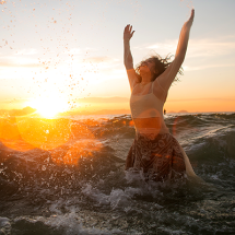 Mulher está no mar, com a água chegando à cintura, de braços erguidos em gesto de celebração. Ela usa blusa clara e saia estampada, enquanto ondas se quebram ao redor. Ao fundo, o sol se põe no horizonte, criando luz dourada e reflexos na água, transmitindo sensação de liberdade, alegria e conexão com a natureza.