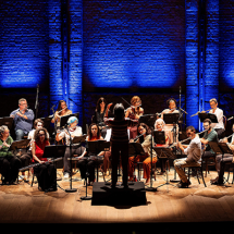 A Orquestra de Flautas da Casa do Choro se apresenta em palco, com vários músicos sentados em semicírculo, tocando diferentes tipos de flauta. Ao centro, a regente está de costas para o público, conduzindo o grupo. O palco é de madeira e o fundo de parede de tijolos é iluminado por luz azul, criando um ambiente solene e elegante, que valoriza a apresentação musical coletiva.