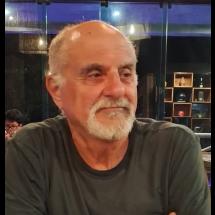 Retrato do professor Alberto Cipiniuk, um homem branco, de aparência madura, com cabelos curtos e grisalhos e barba branca bem aparada. Ele veste camiseta escura e está sentado, com os braços apoiados à frente do corpo. Seu rosto está levemente voltado para a direita, com expressão serena e olhar atento, como se estivesse ouvindo alguém. Ao fundo, o ambiente é interno, com iluminação quente e estantes com objetos decorativos desfocados, sugerindo um espaço cultural ou acadêmico.