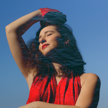 A imagem mostra a cantora e pianista Julie Wein em um retrato artístico. Mulher jovem, de pele clara, fotografada de baixo para cima contra um céu azul. Ela tem cabelos longos e escuros, soltos e levemente ondulados. Usa um vestido vermelho sem mangas e levanta um dos braços acima da cabeça. Seus olhos estão fechados, com expressão serena. Um efeito visual sobreposto lembra ondas de água, criando clima poético e sensorial.