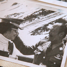 Imagem apresentada no documentário O pacto de Adriana. Trata-se de uma fotografia antiga em preto e branco colada em página de álbum, com borda clara e textura levemente amarelada, marcada por discretos riscos diagonais. Na foto, uma jovem de boné militar e jardineira olha para um homem fardado com medalhas, que toca seu rosto em gesto aparentemente afetuoso. Ao fundo, há uma paisagem com água e montanhas. O enquadramento do álbum reforça o caráter de memória e arquivo histórico.