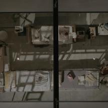 Imagem vista de cima de uma cela coletiva, mostrada como cena do documentário Torre das Donzelas: Memórias e Resistências de Presas Políticas da Ditadura Militar. O espaço é dividido por grades e sombras projetadas, com colchões finos no chão, cobertores dobrados e poucos objetos pessoais espalhados, como livros, roupas e recipientes. A composição rígida e simétrica transmite sensação de confinamento, vigilância e precariedade, reforçando o contexto de repressão e resistência vivido pelas presas políticas.