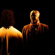 Fotografia de cena teatral da peça “Abdias do Nascimento”. Em palco escuro, dois atores estão frente a frente sob iluminação quente e concentrada. À direita, o ator Lincoln Oliveira aparece de frente, usando terno escuro, óculos, lenço no pescoço e um chapéu de inspiração africana. À esquerda, de costas para a câmera, está o ator Fernando Porto, com cabelos em dreadlocks e camisa clara. O fundo preto destaca o encontro dramático entre os personagens.