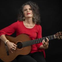 Fotografia de retrato da cantora e compositora Anna Paes. Ela é uma mulher de pele clara, com cabelos cacheados grisalhos, usando blusa vermelha brincos dourados e batom vermelho. Sentada, segura um violão e posiciona os dedos nas cordas, como se estivesse tocando. Olha levemente para cima com expressão serena e confiante. O fundo é preto, destacando a artista e o instrumento.