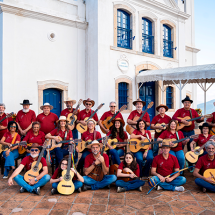 A imagem mostra a Orquestra Caipirando reunida ao ar livre para uma foto coletiva. O grupo é formado por várias pessoas vestidas com camisas vermelhas e chapéus, muitas segurando violas e violões. Alguns integrantes estão sentados no chão ou em bancos, enquanto outros permanecem em pé atrás. Ao fundo, vê-se um edifício histórico de paredes brancas e janelas azuis, com uma área coberta ao lado. O ambiente ensolarado e descontraído destaca o caráter coletivo e musical do grupo.
