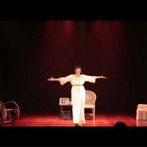 Fotografia de cena teatral com iluminação avermelhada. No centro do palco, a atriz Luciana Borghi está em pé com os braços abertos, interpretando a peça “Na Casa do Rio Vermelho: O Amor de Zélia e Jorge”. Ela veste um vestido longo branco com detalhes dourados. Ao redor, o cenário inclui uma cadeira de balanço, uma poltrona de palha, uma pequena mesa com objetos e caixas, compondo um ambiente doméstico. O palco tem piso de madeira e fundo escuro.