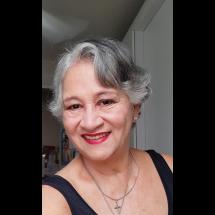Fotografia de retrato da professora e escritora Denise Gals. Ela é uma mulher de pele clara, com cabelos curtos e grisalhos levemente ondulados. Sorri para a câmera, usando batom vermelho e brincos pequenos. A imagem é um close do rosto, com fundo neutro e desfocado, destacando sua expressão acolhedora.