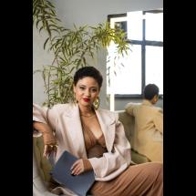 Fotografia de retrato da consultora de imagem e estilo Laís de Souza. Ela é uma mulher negra de pele clara, com cabelos curtos e cacheados. Usa brincos grandes dourados, batom vermelho e maquiagem marcante. Está sentada em um sofá, vestindo blazer claro sobre um top marrom, com uma das mãos apoiada no braço do assento. Olha para a câmera com expressão confiante. Ao fundo há uma planta, uma janela e um espelho que reflete parte do ambiente.
