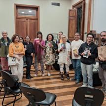 Fotografia de um grupo de cerca de dez pessoas reunidas na sala de leituras do CCJF, durante uma edição anterior do clube de leitura. Os participantes estão lado a lado, sorrindo e olhando para a câmera; alguns seguram livros. O grupo é diverso em idade e aparência. O espaço tem piso de madeira, portas altas de madeira e estantes de livros ao fundo. Em primeiro plano aparecem algumas cadeiras, sugerindo um ambiente de encontro e conversa.