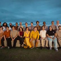 Fotografia de grupo da Orquestra Mil Tons ao ar livre, ao entardecer. Cerca de vinte integrantes estão organizados em duas fileiras — alguns sentados em um banco de pedra e outros em pé atrás. O grupo é diverso em idade, gênero e aparência. Ao fundo, vê-se o mar sob um céu nublado em tons azulados. A iluminação suave destaca os rostos, criando uma atmosfera serena e contemplativa.