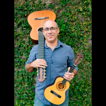 Fotografia de retrato do violonista, cavaquinista, compositor e arranjador Carlos Chaves. Ele é um homem de pele clara, careca, usando óculos e camisa azul de mangas longas. Sorri levemente para a câmera enquanto segura um violão apoiado no ombro e um cavaquinho nas mãos. Ao fundo há uma parede coberta por folhagens verdes, criando um cenário natural que destaca os instrumentos musicais.