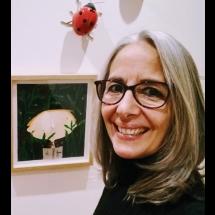 Fotografia de retrato da escritora e artista plástica Patrícia Montês. Ela é uma mulher de pele clara, com cabelos longos e grisalhos, usando óculos de armação escura. Sorri olhando para a câmera. Ao fundo, em uma parede clara, há um quadro emoldurado com uma ilustração de uma luminária cercada por folhas verdes, sugerindo um ambiente de exposição ou galeria.
