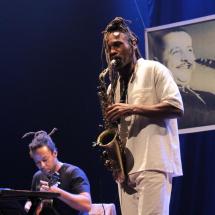 Fotografia de apresentação musical em palco com iluminação azul. Em primeiro plano, o saxofonista Rocino Crispim, homem negro com longos dreadlocks presos no alto da cabeça, toca saxofone vestindo roupa clara. Ao fundo, à esquerda, um dos violonistas da Regional Caboclo aparece sentado, concentrado no violão. Na parte de trás do palco há a projeção de uma foto em preto e branco do músico Luiz Americano tocando saxofone.