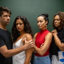 Fotografia da peça teatral O Último Dia. Quatro personagens aparecem lado a lado diante de fundo verde escuro (composição fechada e intensa). À esquerda, um homem segura o braço de uma mulher com expressão séria. As duas mulheres ao centro mantêm os olhos fechados ou baixos, com semblantes tensos e introspectivos. À direita, outra mulher observa com preocupação, mãos unidas junto ao peito. A proximidade dos corpos e as expressões contidas sugerem conflito emocional, luto ou um momento decisivo na narrativa.