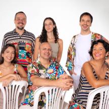 Fotografia de retrato do sexteto Ordinarius. Seis integrantes posam diante de fundo branco, organizados em dois planos: três sentados em cadeiras plásticas à frente e três em pé atrás. O grupo é diverso em gênero e aparência, todos sorrindo e olhando para a câmera. Vestem roupas leves e coloridas, com estampas variadas. A composição é descontraída e destaca a união do grupo musical.