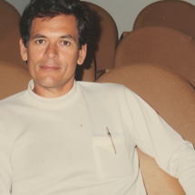 O dramaturgo Roberto Athayde aparece sentado em uma poltrona de auditório, vestindo uma blusa branca de mangas longas com uma caneta no bolso. Ele olha serenamente para a câmera, em meio a fileiras de cadeiras marrons vazias ao fundo, sugerindo um momento de tranquilidade em um ambiente cultural.