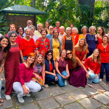 Fotografia de grupo do Coral da Tarde em Canto Plural ao ar livre. Cerca de trinta integrantes estão reunidos em um jardim, entre árvores e plantas, organizados em fileiras — alguns em pé e outros agachados à frente. O grupo é diverso em idade e aparência, vestindo roupas coloridas, com predominância de tons quentes. Todos sorriem e olham para a câmera, transmitindo um clima de alegria e convivência.