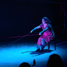Cena da peça Filipa mostra uma mulher sentada em uma cadeira, em palco escuro iluminado por luz azul. Ela veste vestido vermelho e puxa com força um fio ou corda esticada à sua frente, com expressão intensa e concentrada. O cenário é minimalista, com poucos objetos, e linhas finas coloridas atravessam o espaço, criando tensão visual e reforçando o clima dramático da cena.