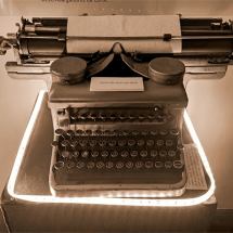 Máquina de escrever antiga em exposição. O objeto está sobre um pedestal iluminado, com papel encaixado no rolo e teclas circulares visíveis. A iluminação destaca os detalhes metálicos e o desgaste do equipamento, sugerindo valor histórico. Ao fundo, há uma parede neutra com pequena placa informativa acima.