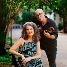 Fotografia de retrato ao ar livre da cantora e violonista Anna Paes e do violonista e cavaquinista Carlos Chaves. Em primeiro plano, Anna Paes, mulher de pele clara com cabelos cacheados grisalhos, está sentada em um banco de praça, sorrindo para a câmera, usando vestido estampado. Ao lado e ligeiramente atrás, Carlos Chaves, homem de pele clara, careca e com óculos, veste roupa escura e segura um violão, com postura de quem toca. O fundo mostra árvores e um caminho, criando um ambiente natural e acolhedor.