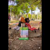 A imagem mostra um parque com árvores e folhas secas espalhadas pelo chão. Em primeiro plano, de frente para a câmera, há um livro com título PÉDEPOESIA apoiado em duas pedras, sobre o chão. Há duas bonecas de pano sentadas nas pedras, de costas uma para a outra. Uma das bonecas tem cabelos escuros e usa vestido vermelho; a outra tem cabelos claros e veste roupas em tons neutros. Ao fundo, há vegetação e um caminho sombreado, criando um ambiente natural e tranquilo.