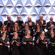 Apresentação de coral em palco mostra grupo numeroso de cantores organizados em fileiras. Todos vestem roupas formais em tons escuros, com alguns homens usando gravatas vermelhas. Cada integrante segura uma partitura e canta com expressão concentrada. Ao fundo, um painel geométrico branco cria textura visual. A iluminação é clara e uniforme, destacando o conjunto e a harmonia da apresentação.