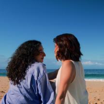 Cena do filme Azul mostra duas mulheres sentadas na areia de uma praia, próximas ao mar. Elas estão de perfil, voltadas uma para a outra, com expressões suaves e íntimas. Uma veste camisa azul e tem cabelos longos e cacheados; a outra usa blusa clara e tem cabelo curto. Ao fundo, o mar azul e o céu aberto criam um clima tranquilo e contemplativo.