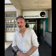 Retrato do professor Erick Felinto em área externa do Instituto de Artes da UERJ. Ele aparece em primeiro plano, com cabelos grisalhos e barba curta, usando camisa clara de linho e olhando para a câmera com leve sorriso. Ao fundo, vê-se a entrada do instituto, com a placa “Instituto de Artes” e parte do corredor interno do prédio.