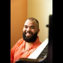 Retrato do pianista Thalyson Rodrigues sorrindo para a câmera. Ele aparece em primeiro plano, com barba cheia e camisa em tom alaranjado. À direita da imagem, parte desfocada de um piano sugere o contexto musical do ambiente. O fundo em tons quentes reforça a atmosfera acolhedora da cena.