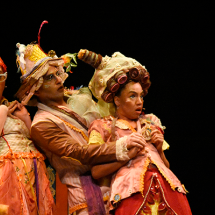 Cena da peça teatral “Era Uma Vez um Tirano”. Três atores aparecem juntos no palco, usando figurinos coloridos e extravagantes em tons de laranja, rosa e dourado, com maquiagem e acessórios exagerados. As personagens fazem expressões de surpresa e comicidade, inclinando os corpos para trás em movimento teatral. O fundo escuro destaca o clima lúdico e fantasioso da encenação.