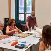 Grupo de pessoas reunidas em torno de uma mesa durante atividade ou oficina em sala interna. Um homem de barba grisalha está em pé, distribuindo folhas de papel, enquanto outras pessoas sentadas organizam materiais e fazem anotações. Sobre a mesa há cadernos, garrafas de água, estojos e papéis. Ao fundo, uma janela alta com moldura verde ilumina o ambiente de trabalho coletivo.