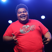 O comediante Big Jaum se apresenta no palco com expressão alegre e sorriso aberto. Vestindo camiseta vermelha e segurando um celular, ele faz um gesto com uma das mãos enquanto interage com o público. A iluminação azul ao fundo cria contraste com a roupa e reforça a atmosfera vibrante do show.