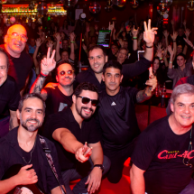 Integrantes da banda Cadillac posam com a plateia ao fundo durante apresentação. Reunidos em frente ao palco, os músicos sorriem e fazem gestos para a câmera, enquanto o público lota a casa de shows com os braços erguidos. A iluminação vermelha e o ambiente festivo destacam a energia do espetáculo.