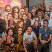 Integrantes da equipe do FID:RIO (Festival Internacional de Cinema de Não Ficção do Rio de Janeiro) posam para fotografia coletiva. Reunidos diante do painel de divulgação do festival, cerca de vinte pessoas sorriem para a câmera em clima de celebração. Ao fundo, a identidade visual do evento reforça o contexto cinematográfico e colaborativo da imagem.