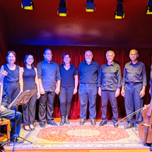 Integrantes do Coro Madrigal da ACC posam em fotografia. O grupo está alinhado diante de uma cortina vermelha, vestindo trajes pretos. Nas extremidades, dois músicos seguram instrumentos de cordas antigos, um alaúde e uma viola da gamba. A iluminação cênica em tons quentes destaca os participantes e o tapete ornamentado que compõe o cenário, criando atmosfera de concerto de música vocal e de câmara.