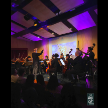 Apresentação da Orquestra Filarmônica Fluminense em palco iluminado por luzes em tons de roxo e azul. Ao centro, o maestro rege os músicos com os braços erguidos. A orquestra é formada por instrumentistas de cordas, incluindo violinos, violoncelos e contrabaixos, dispostos diante do público. Ao fundo, uma tela de projeção exibe a identidade visual do concerto. O ambiente destaca a execução coletiva e a atmosfera solene da apresentação.