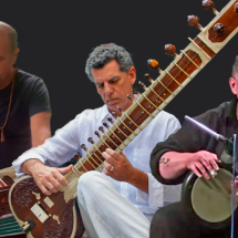 Integrantes do Trio Shiva Gita (Índia) em montagem promocional. Ao centro, um músico toca uma sitar, instrumento tradicional indiano de cordas. À esquerda, outro integrante toca um harmonium, teclado de fole utilizado na música clássica e devocional da Índia. À direita, um percussionista executa um tambor de mão. Os três aparecem sobre fundo escuro, destacando os instrumentos e a diversidade sonora do grupo.