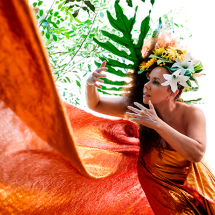 Retrato da cantora Zilá Lima em cenário cercado por elementos da natureza. Com vestido em tons de laranja e adorno floral na cabeça, ela faz gestos expressivos com as mãos enquanto olha para a frente. Grandes folhas verdes e um tecido alaranjado em movimento compõem a cena, criando uma atmosfera vibrante e inspirada na natureza.