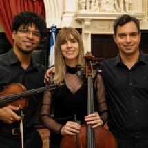 Integrantes do Trio Movimento Musical posam lado a lado em um salão de arquitetura clássica. À esquerda, o violinista Luís Felipe Oliveira segura um violino e o arco. Ao centro, a violoncelista Denise Emmer segura um violoncelo apoiado à frente do corpo. À direita, o pianista João Paulo Romeo sorri para a câmera. Os três vestem roupas pretas e têm ao fundo detalhes em madeira escura, cortinas vermelhas e elementos ornamentados do ambiente.