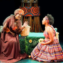 Cena do espetáculo A Bela Adormecida. Em um palco com fundo escuro, uma senhora negra ajoelhada veste longo traje marrom com adornos em tons terrosos, turbante e maquiagem cênica. Ela gesticula enquanto fala com uma jovem negra sentada sobre as pernas à sua frente, que usa vestido amplo e estampado em cores vibrantes, com faixa colorida nos cabelos, e a observa atentamente. Ao lado da senhora há um cesto de vime. Ao fundo, um elemento cenográfico com estampas geométricas africanas completa a composição.
