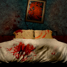 Still do filme Bora!. Um quarto escuro é dominado por uma cama de casal com lençóis brancos marcados por manchas vermelhas que lembram sangue e por marcas de mãos espalhadas sobre o tecido. Ao fundo, a parede apresenta rachaduras, respingos avermelhados e um quadro torto com uma pintura abstrata em tons de vermelho e preto. A iluminação baixa e o ambiente desordenado reforçam a atmosfera de tensão e mistério.