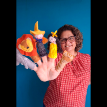 A escritora Luciana Belém sorri para a câmera enquanto exibe dedoches de personagens infantis colocados nos dedos de uma das mãos: um elefante, um leão, um menino, uma menina e um tigre. Ao fundo, um painel azul destaca a cena. A escritora usa óculos, blusa estampada em tons de vermelho e mantém a mão estendida em direção à câmera, aproximando os dedoches do primeiro plano.
