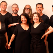 Integrantes da equipe da peça Café, Almoço e Jantar posam reunidos para a fotografia. O grupo é formado por oito pessoas, distribuídas em duas fileiras, vestindo roupas predominantemente pretas. Em primeiro plano, duas mulheres sorriem abraçadas, enquanto os demais integrantes se posicionam ao redor, alguns com as mãos apoiadas nos ombros dos colegas. O fundo claro e as expressões descontraídas reforçam o clima de convivência e celebração do grupo.